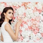 A woman stands gracefully in front of a vibrant flowerwall of flowers, exuding elegance and beauty.