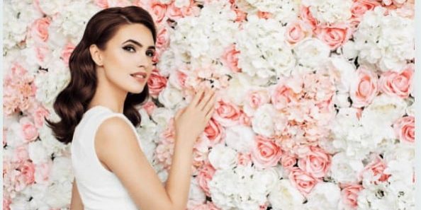 A woman stands gracefully in front of a vibrant flowerwall of flowers, exuding elegance and beauty.