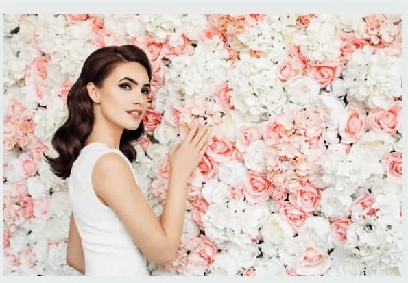 A woman stands gracefully in front of a vibrant flowerwall of flowers, exuding elegance and beauty.